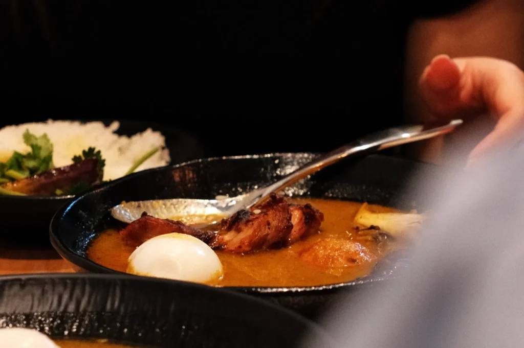 A bowl of curry with meat, boiled eggs, and a spoon, accompanied by a plate of rice and salad in the background.