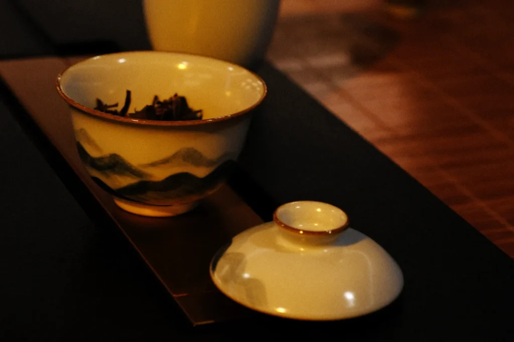 Warmly lit yellow ceramic tea bowl and matching lid rest on a dark tray, with painted landscape details and a calm, intimate tea setting.