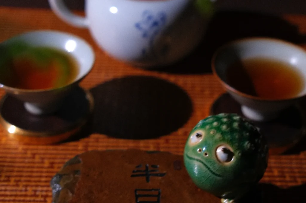 A green, frog-like figurine is positioned in front of two small tea cups and a teapot, with a stone inscribed with Chinese characters in the foreground, all set on a bamboo mat.