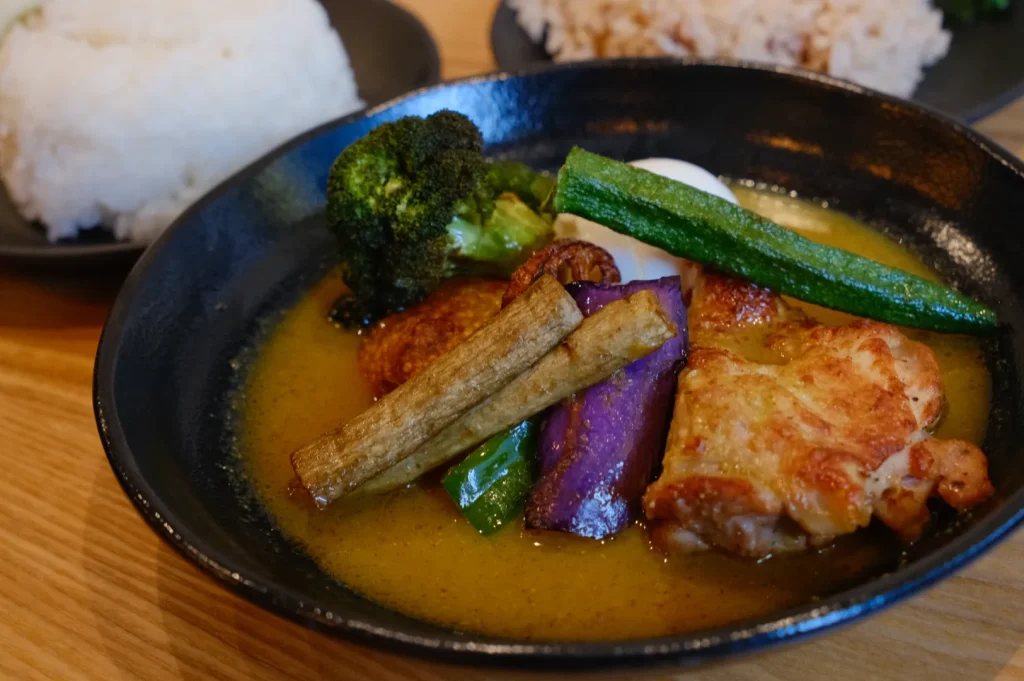 A bowl of curry with chicken, broccoli, okra, and various vegetables, served with rice on the side.