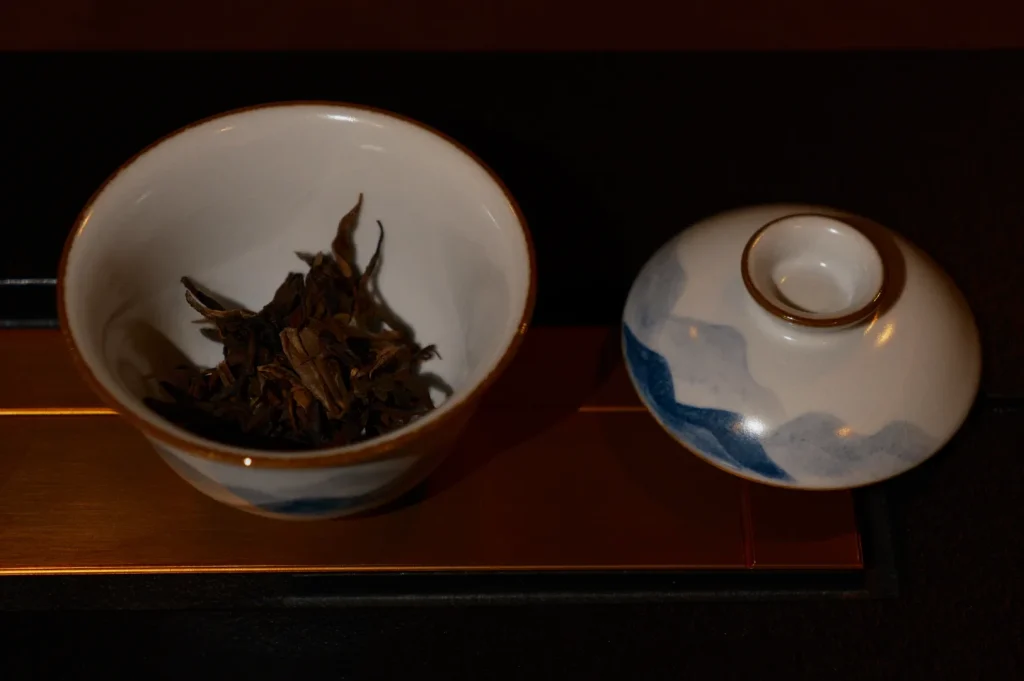 A white ceramic bowl filled with dried tea leaves sits beside a lidded blue-and-white tea bowl on a dark wooden tray, creating a calm mood.