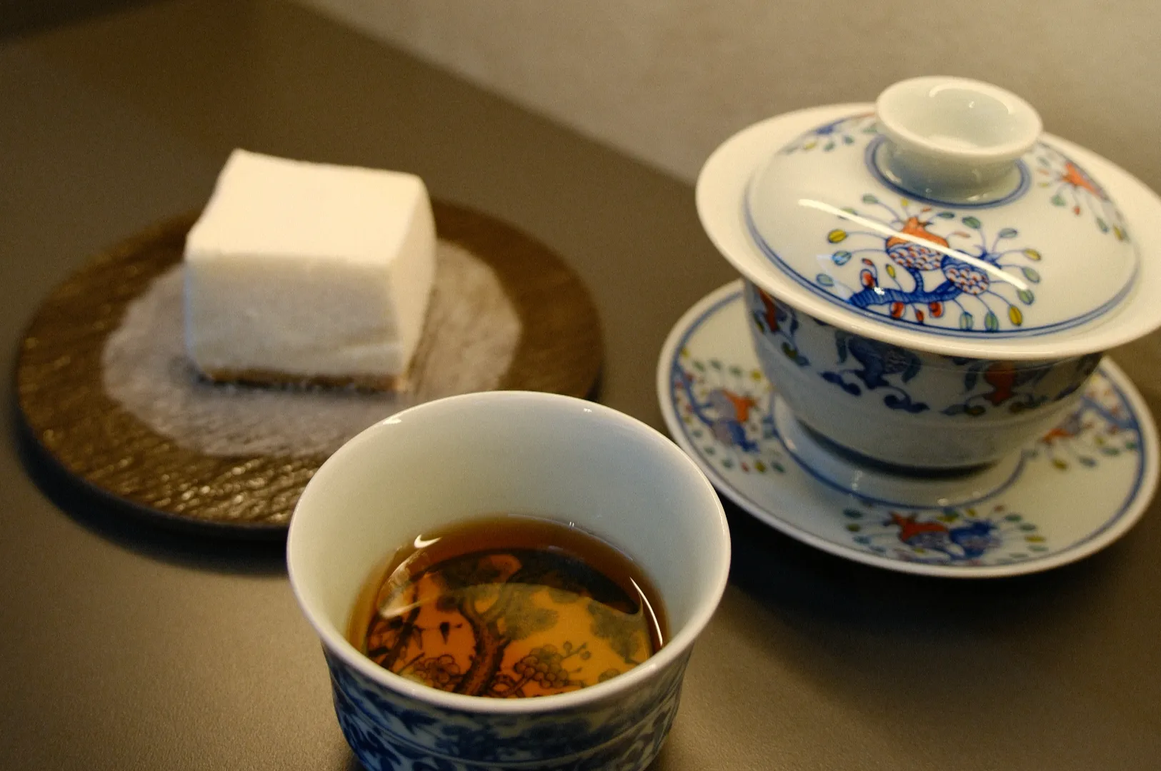 A cup of amber tea sits beside a blue-and-white floral lidded gaiwan and a square of white cake on a dark table, creating a calm tea-time scene.