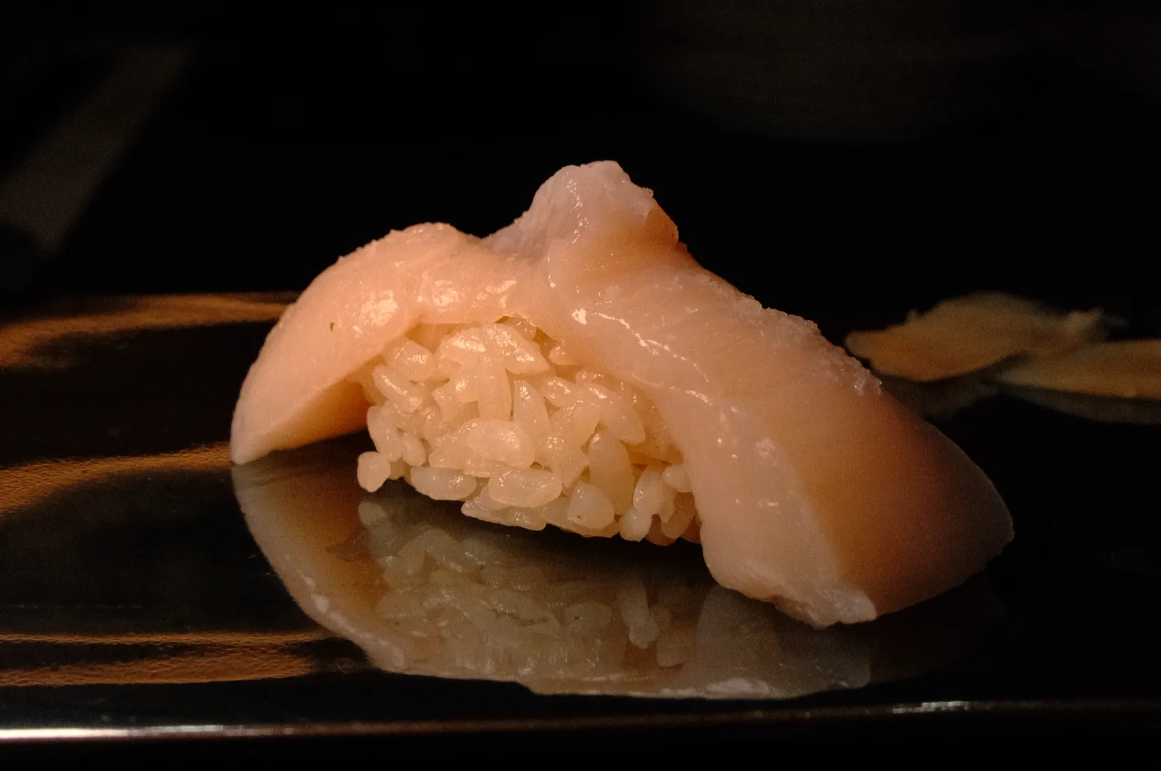 Close-up of pale, translucent fish nigiri sushi draped over a compact mound of rice on a glossy black plate, creating a simple, elegant mood.