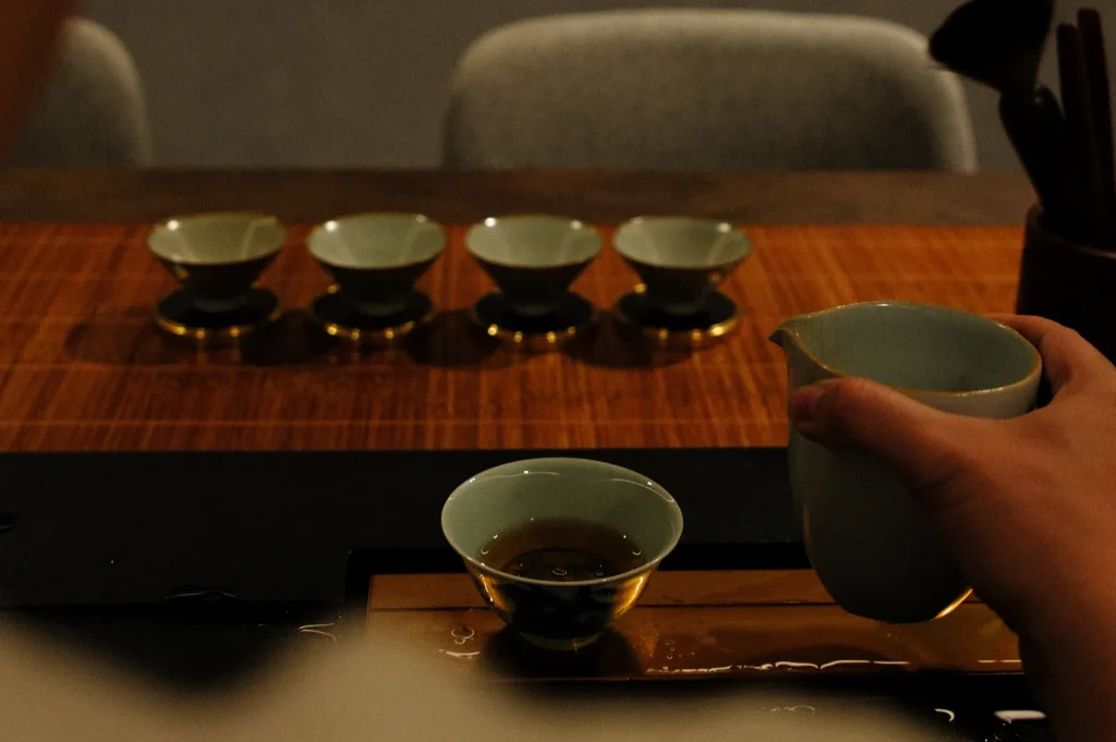 A hand pours tea from a gray-green pitcher into a small cup on a wooden tea table, with four matching cups lined up behind it in warm, quiet light.