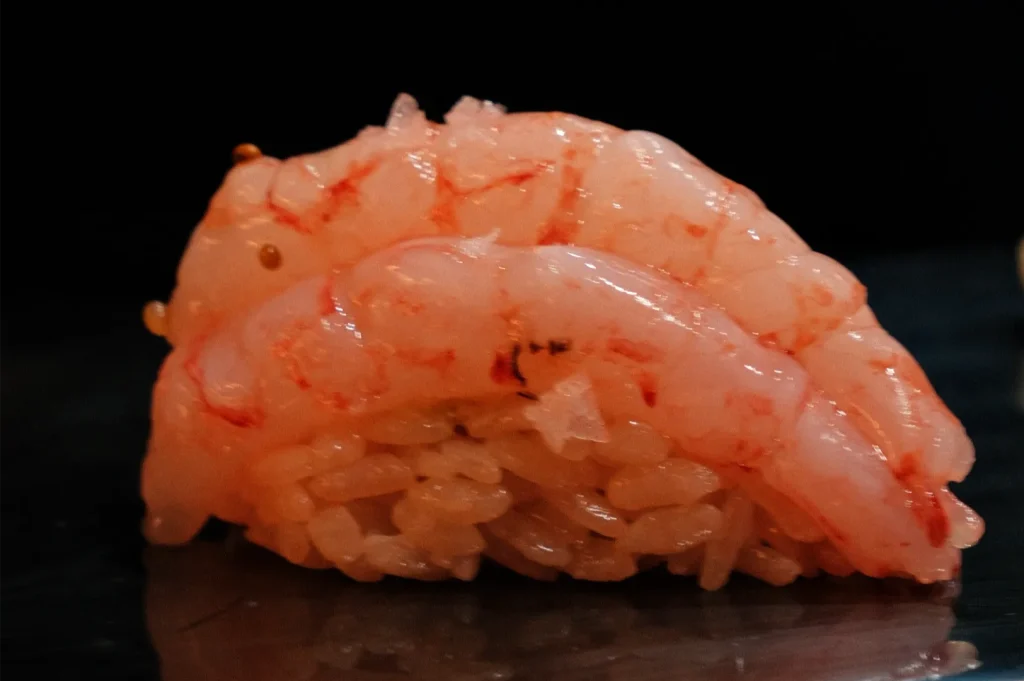 Close-up of glossy pink shrimp nigiri layered over compact rice on a dark surface, isolated against a black background for a dramatic presentation.