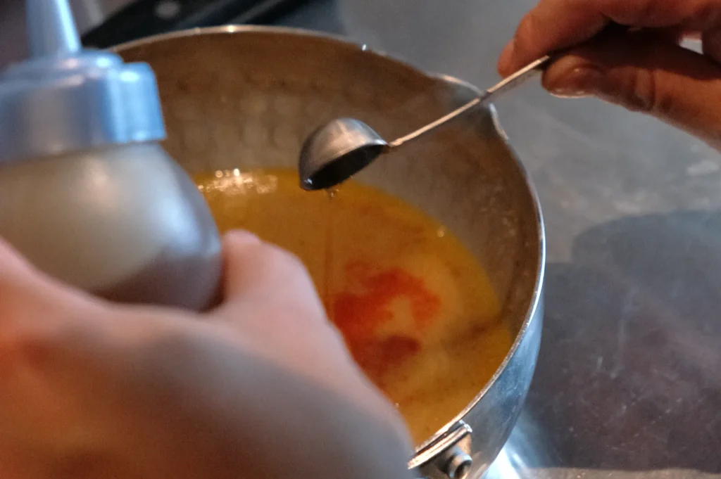 Hands squeeze a bottle and pour liquid from a spoon into a pot of orange-red soup, capturing an intimate, hands-on moment of cooking.