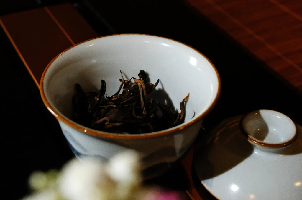A white ceramic tea bowl holds dark loose tea leaves, with its matching lid beside it on a wooden table, creating a warm, quiet mood.