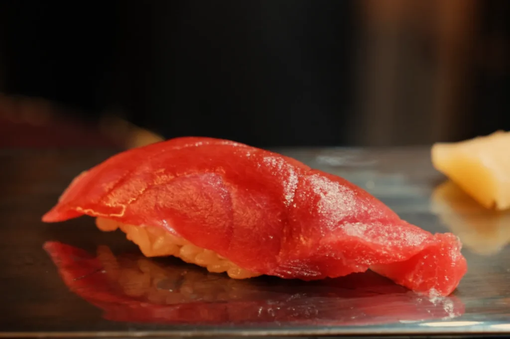 A glossy red tuna nigiri sushi rests on a dark plate, with a pale yellow pickled ginger slice in the background; warm lighting gives the scene an elegant, appetizing feel.