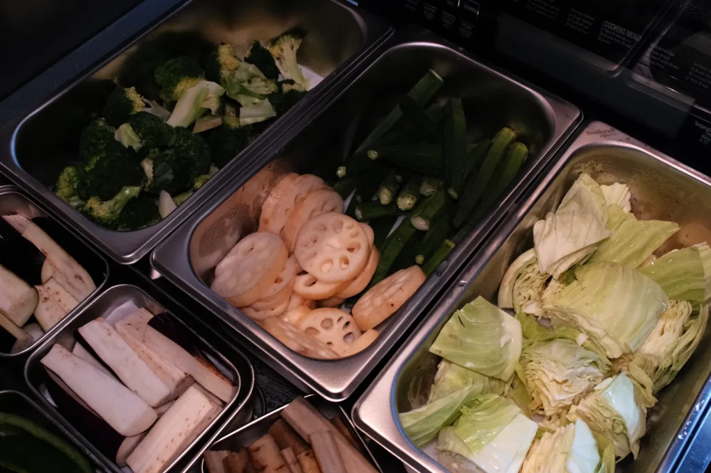 A dimly lit buffet displays metal trays of broccoli, okra, sliced lotus root and carrots, chopped cabbage, and pale mushrooms, ready to serve.