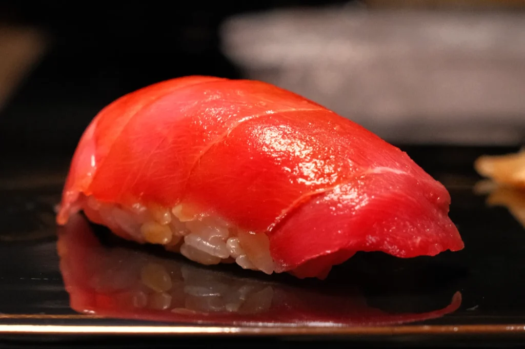 Glossy red tuna nigiri sushi rests on a black plate, its fresh fish draped over compact rice; warm lighting gives the close-up an appetizing feel.
