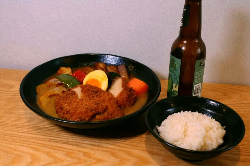 A pairing of the Crunchy Boneless Chicken Thigh soup curry bowl with mixed vegetables served alongside white rice and a bottled craft beer, at Craft Beer by Ki-setsu.