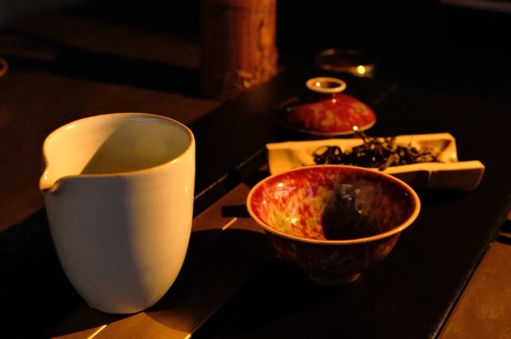 A white ceramic pitcher, mottled red-brown bowl, small lidded pot, and rectangular dish sit on a dark table, warmly lit in a quiet still life.