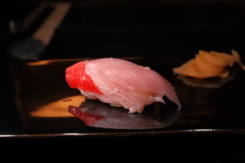 A piece of glossy pink-and-red nigiri sushi rests on a dark plate, with pickled ginger in the background; dramatic lighting creates an elegant, appetizing mood.