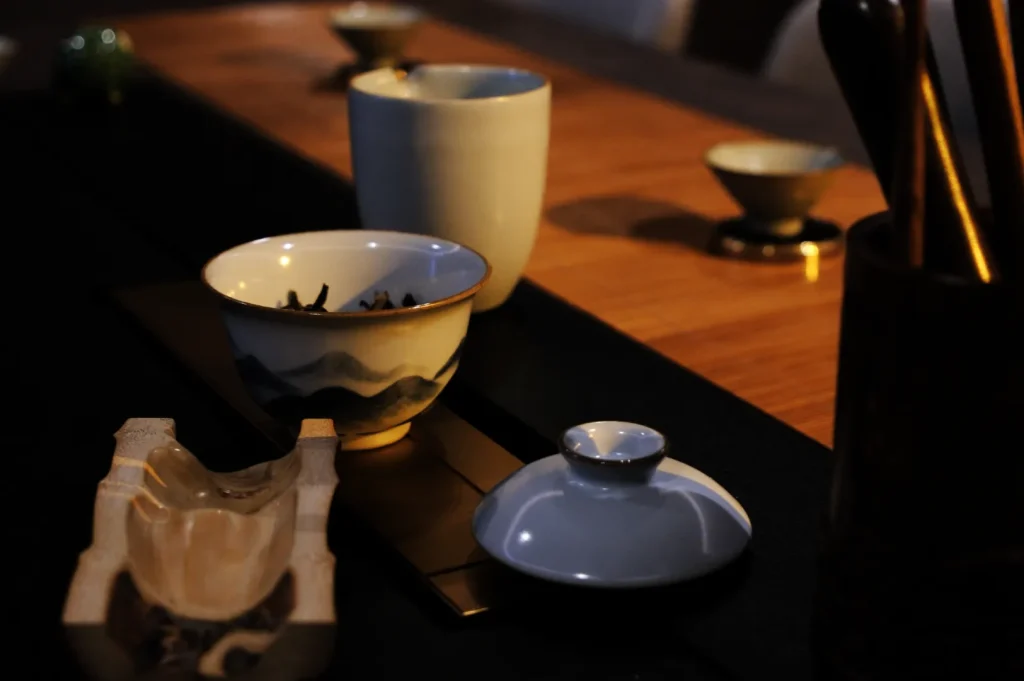 A warmly lit tea setup features a patterned porcelain cup, matching lid, tall ceramic mug, and wooden utensils on a dark tabletop, creating a quiet, intimate mood.