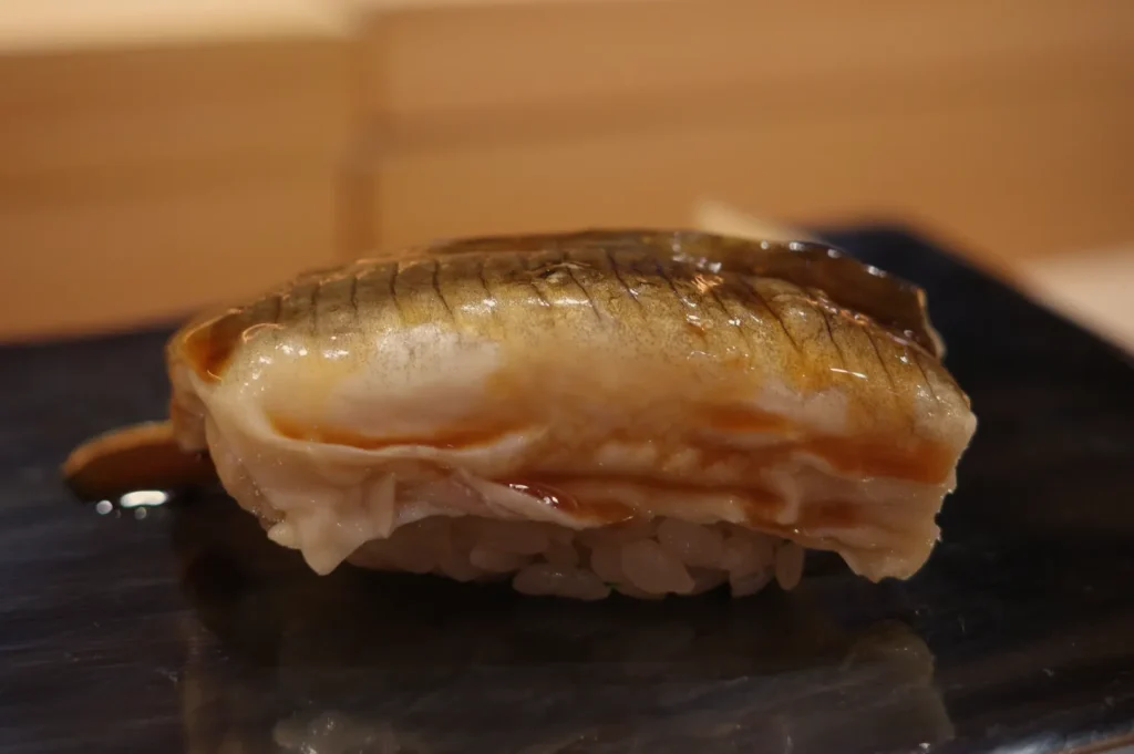 Close-up of a glossy, lightly seared fish nigiri sushi on a black plate, its golden-brown topping resting on compact white rice in warm light.