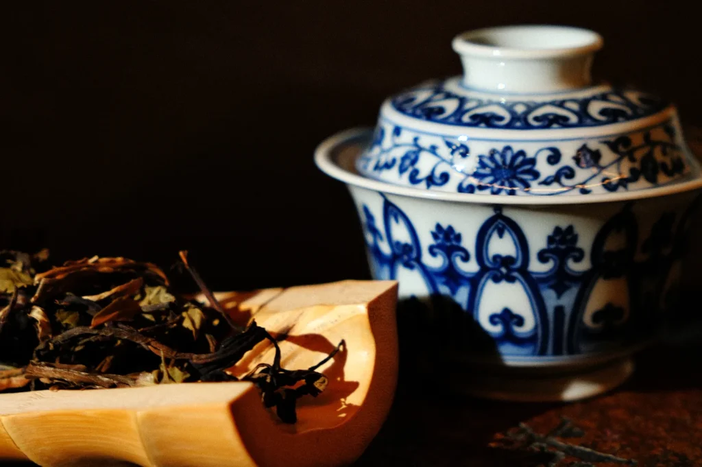 Loose tea leaves sit in a wooden scoop beside an ornate blue-and-white porcelain lidded cup, warmly lit against a dark backdrop, evoking calm.