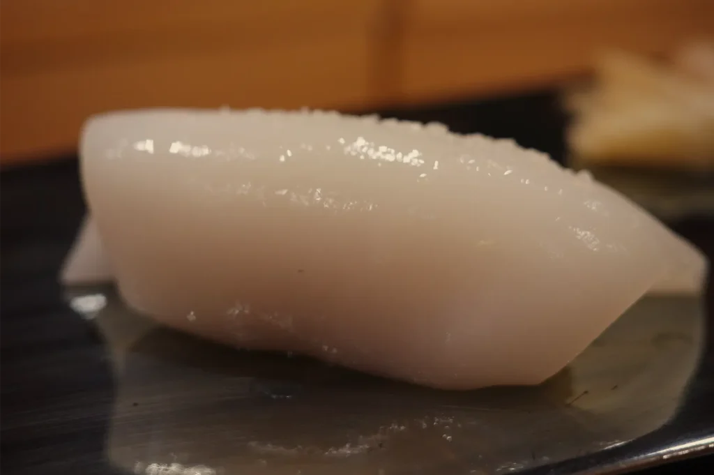 A glossy slice of white fish, likely sashimi, rests on a dark ceramic plate against a warm, softly blurred background, creating a calm, appetizing mood.