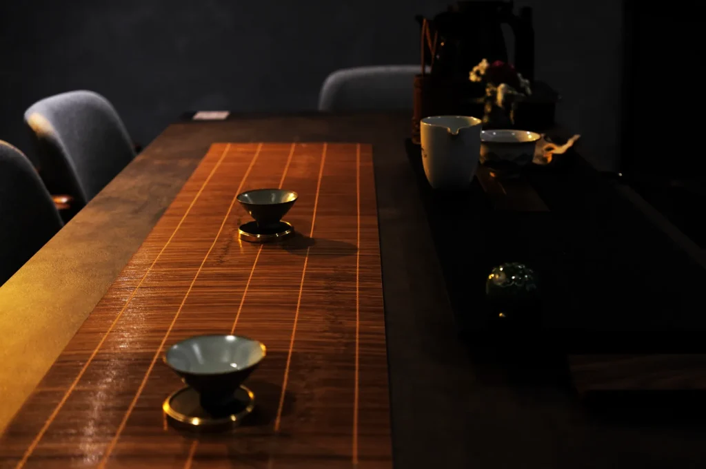 A long wooden tea table displays small ceramic cups and tea vessels in a dim room, creating a quiet, intimate atmosphere.