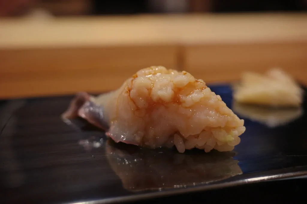 Close-up of glossy pale fish nigiri draped over rice on a black plate, with another piece blurred behind; an elegant, appetizing sushi-bar scene.