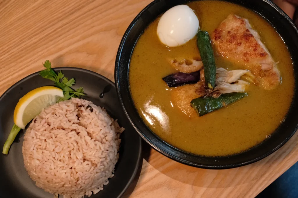 A black bowl of golden curry with a soft egg, browned chicken, and vegetables sits beside molded rice garnished with lemon and herbs on a wooden table.